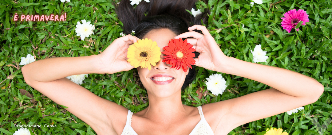 Mulher sorrindo deitada na grama com duas flores cobrindo os olhos